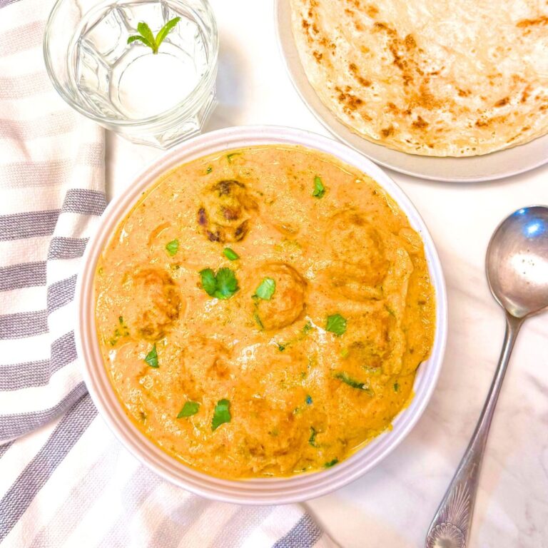 white bowl with mixed vegetable kofta curry placed on a marble along with a napkin, spoon, parathas and a glass of water.