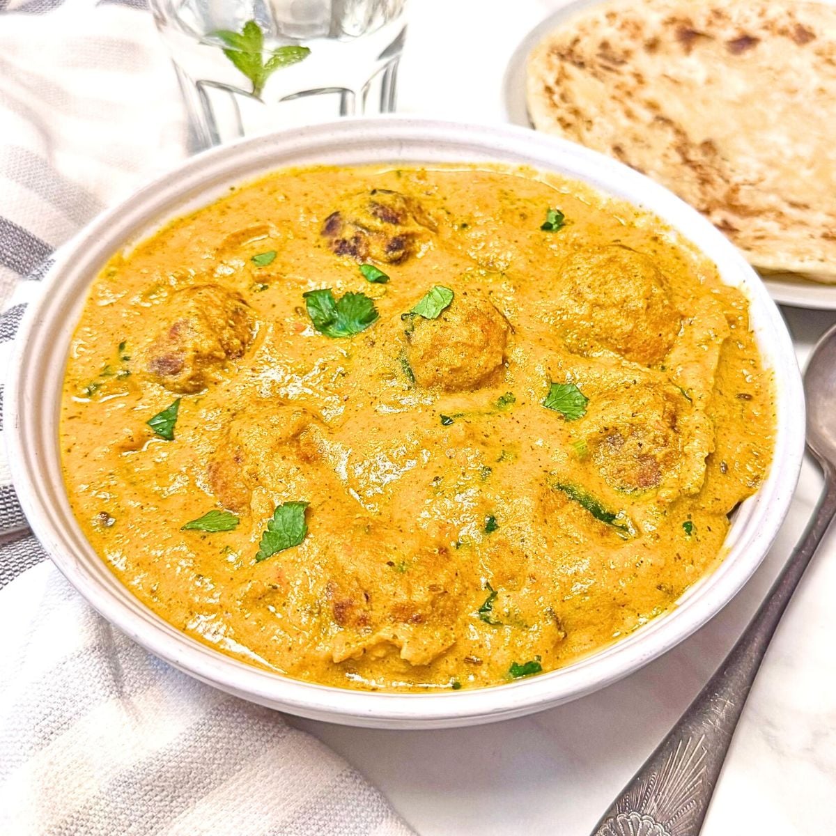 close up picture of mixed vegetable kofta curry in a white bowl placed on marble along with a spoon and plate of parathas.