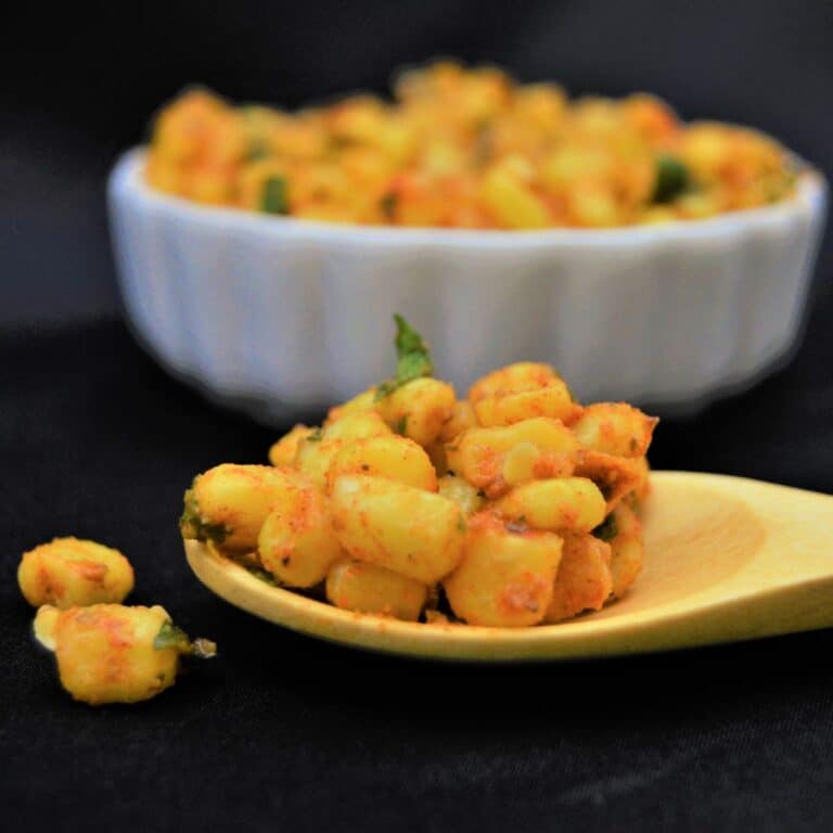a wooden spoon filled with a masala sweet corn placed on a black cloth along with a white bowl of masala corn behind it.