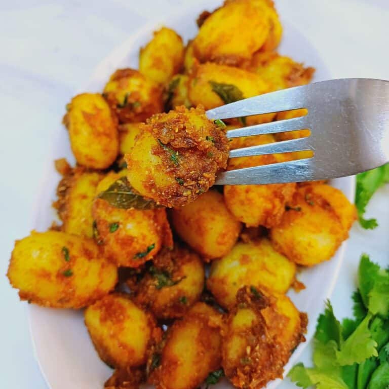 holding a spiced baby potato with a fork on top of a plate of spicy baby potato fry.