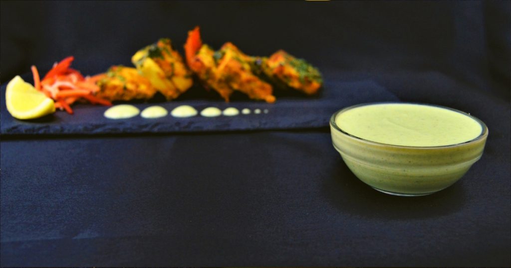 yogurt mint sauce in a small bowl on black cloth with paneer tikka behind it.