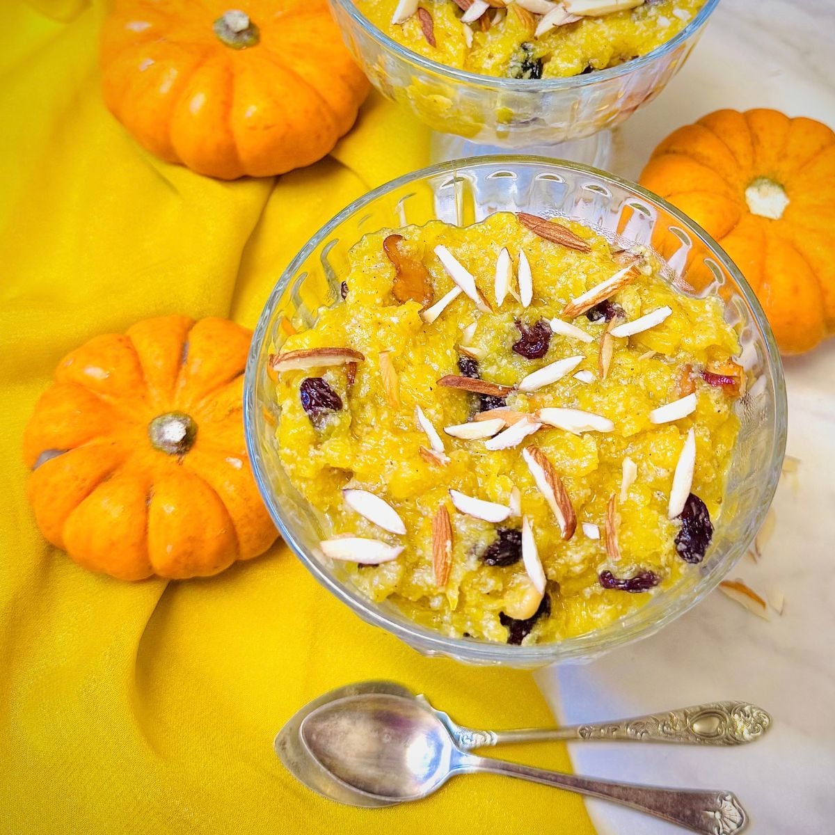 two glass bowls of pumpkin halwa topped with nuts placed on marble along with small pumpkins and a cloth.