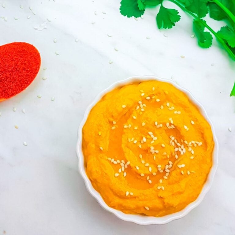 Top view image of carrot dip in a white bowl topped with sesame seeds placed on marble along with a bunch of coriander leaves, wooden spoon of chilli powder.