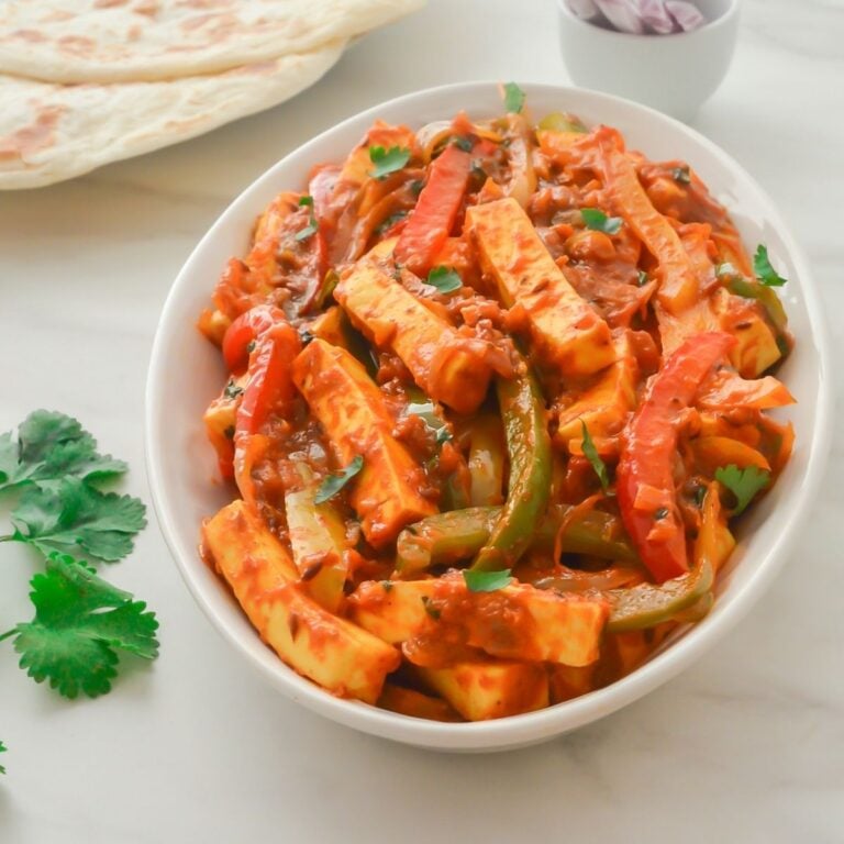 paneer jalfrezi in a white bowl with a paratha, sprig of coriander and onion slices on a marble.