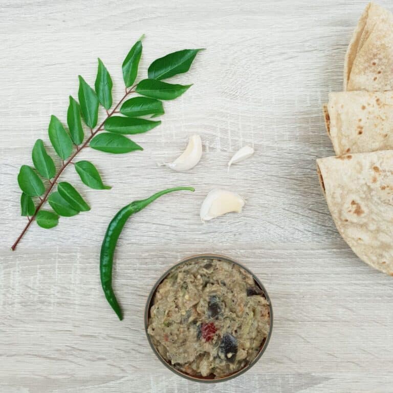 vegan eggplant chutney placed on a wooden table along with green chilli, curry leaves and chapatis.