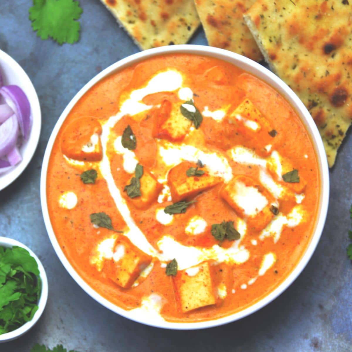 paneer makhani in a white bowl placed on a baking sheet along with a bowl of onion slices, coriander and naan bread.