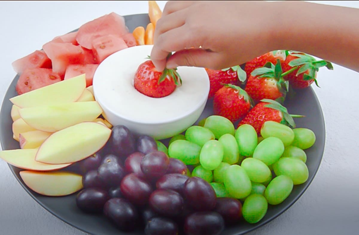 dipping strawberry in fruit yogurt dip placed in the middle of the fruits in a plate.