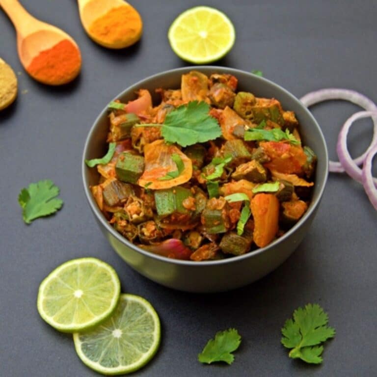 bhindi masala in a black bowl placed on black table along with lemon slices, onion rings and spices.