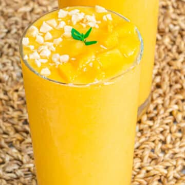 banana pineapple mango smoothie in a glass topped with nuts placed on a mat.