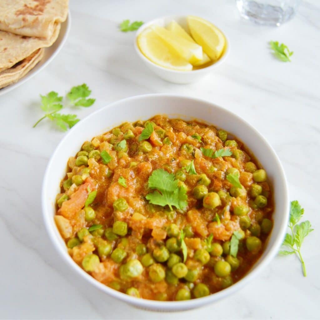 green peas curry in a white bowl placed on a marble along with a pinchbowl of lemon wedges and a plate of chapatis.