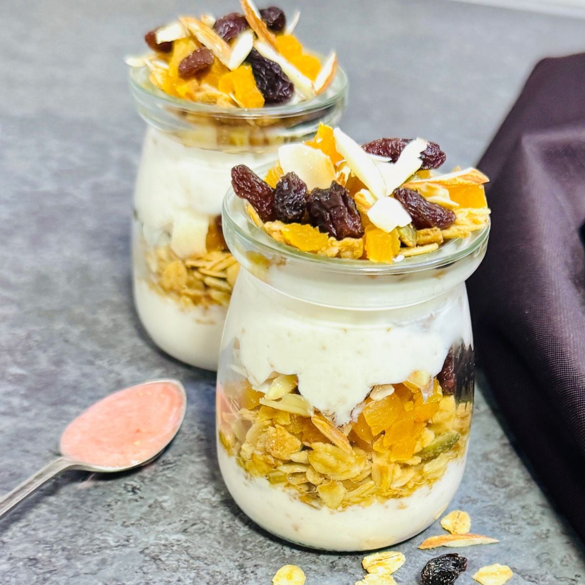 two jars of yogurt granola parfait placed on granite along with a spoon of strawberry honey and a brown cloth.