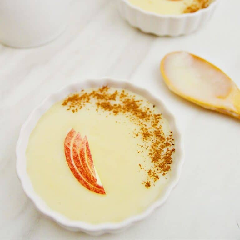 dairy free coconut custard in a white bowl garnished with apple slices placed on a marble along with a wooden spoon.