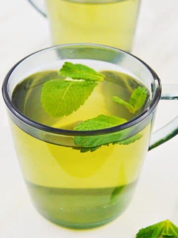 2 glasses of fresh mint tea placed on a white table along with a few mint leaves next to it.