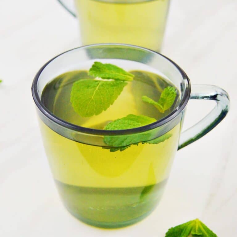 2 glasses of fresh mint tea placed on a white table along with a few mint leaves next to it.
