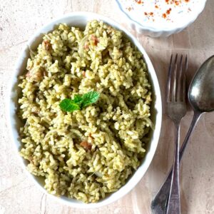 oval plate filled with mint pulao placed on a granite along with cutlery and a bowl of raita.