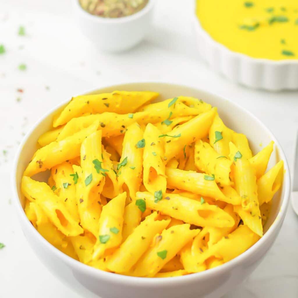 creamy carrot pasta in a white bowl placed on a marblw along with a bowl of carrot sauce and pinch bowl of spice mix.