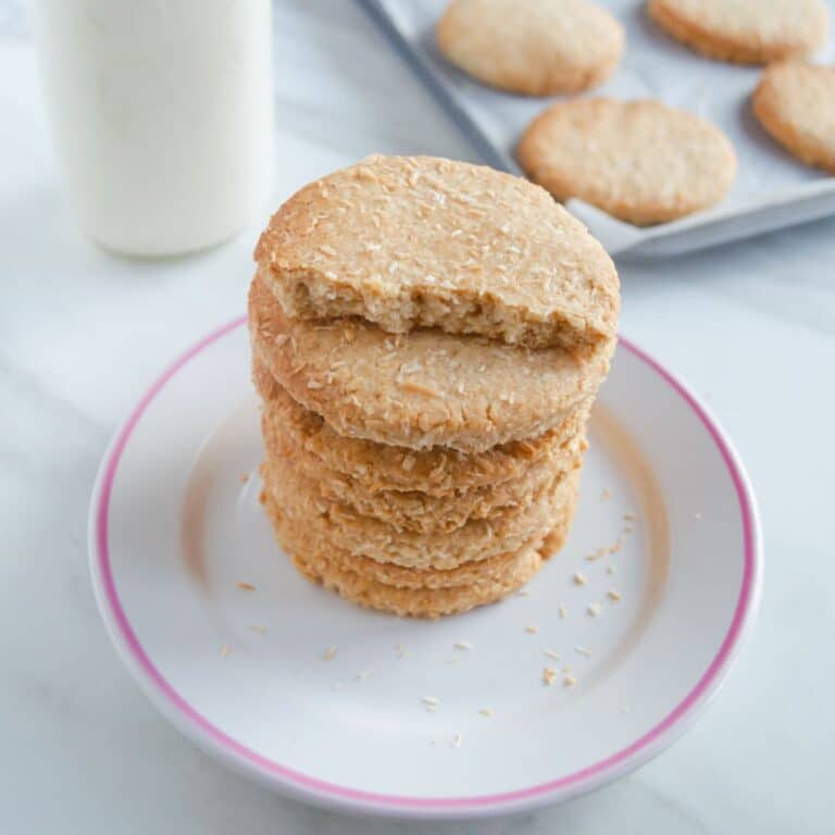 a white plate of stacked eggless coconut biscuits placed on a marble along with a tray of biscuits and milk bottle behind it,