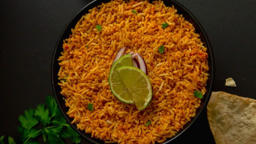 tomato rice in a black bowl with onion slice and half cut lemons on top placed on black board.