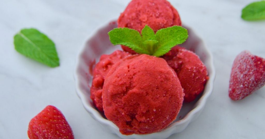 close up shot of scoops of strawberry frozen yogurt with mint leaves on top in a white bowl on marble.