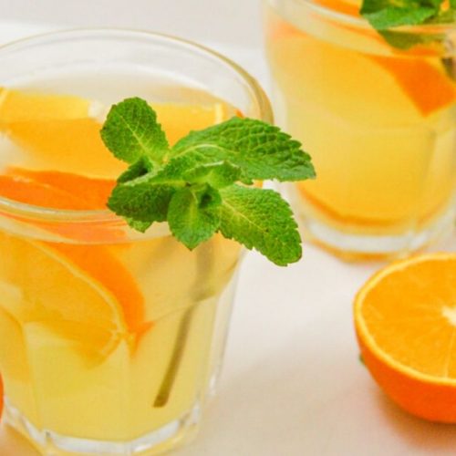 orange syrup in 2 glasses with mint sprig and half cut oranges on a marble.