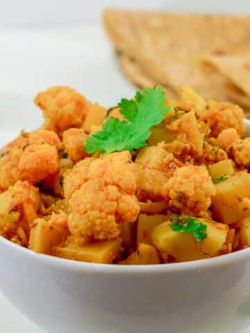 dry aloo gobi in a white bowl placed on a marble along with a plate of chapatis behind it.