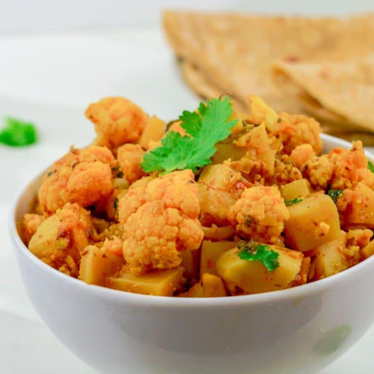 dry aloo gobi in a white bowl placed on a marble along with a plate of chapatis behind it.