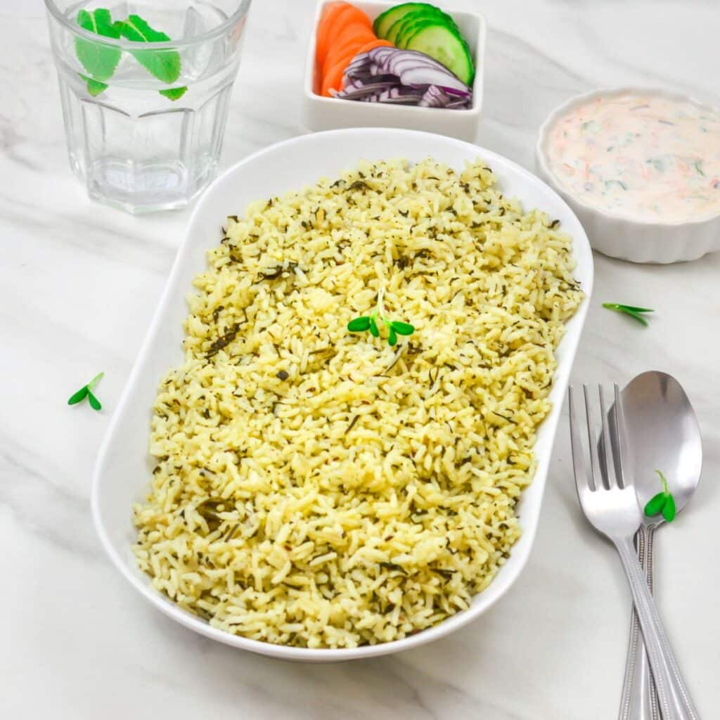 Pin Image of Methi Pulao in a white bowl on a marble background with cutlery, bowl of raita, salad and glass of water.