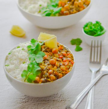white bowl filled with white rice, chickpea and spinach curry with coriander and lemon wedge placed on white table along with 2 forks, pinch bowl of coriander and 2nd bowl of rice and curry behind.