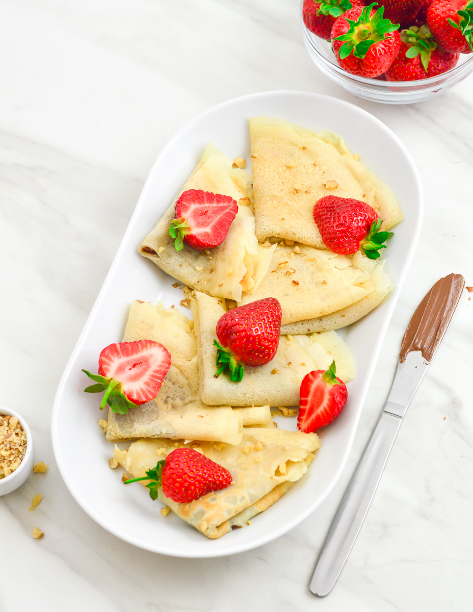 folded nutella crepes topped with halved strawberries arranged in an oval plate placed on a marble along with a knife coated with nutella, bowl of strawberries and a pinch bowl of walnuts.