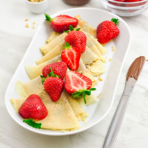 sweet crepes arranged in a white plate with strawberries on top and a knife with chocolate placed on marble.