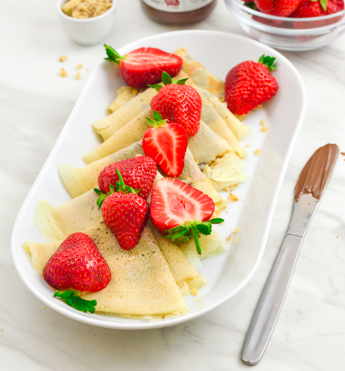 sweet crepes arranged in a white plate with strawberries on top and a knife with chocolate placed on marble.