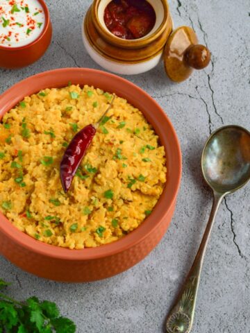 khichdi in a brown bowl placed on a granite along with twigs of coriander, serving spoon, a bowl of yogurt and bowl of pickle.