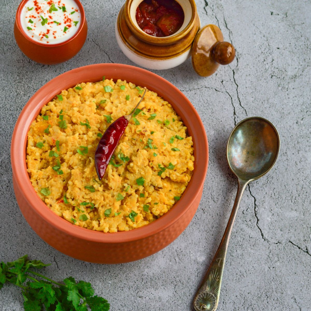 khichdi in a brown bowl placed on a granite along with twigs of coriander, serving spoon, a bowl of yogurt and bowl of pickle.