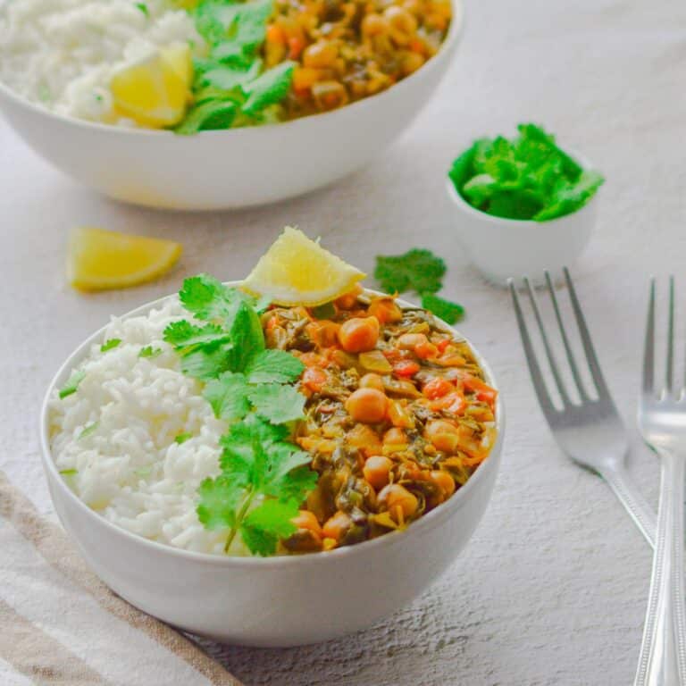 white bowl filled with white rice, chickpea and spinach curry with coriander and lemon wedge placed on white table along with 2 forks, pinch bowl of coriander and 2nd bowl of rice and curry behind.