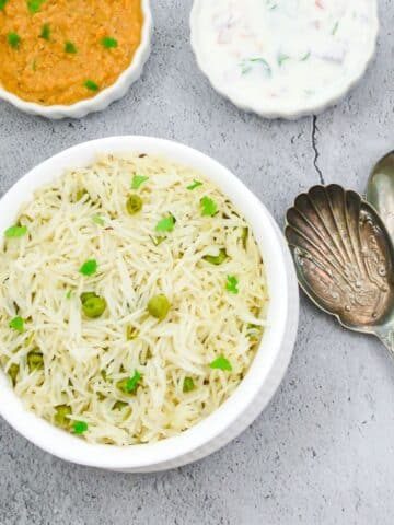 peas pulao in a white bowl and 2 spoons placed on a wooden chopping board along with a bowl of gravy, raita, sliced onion on a granite.