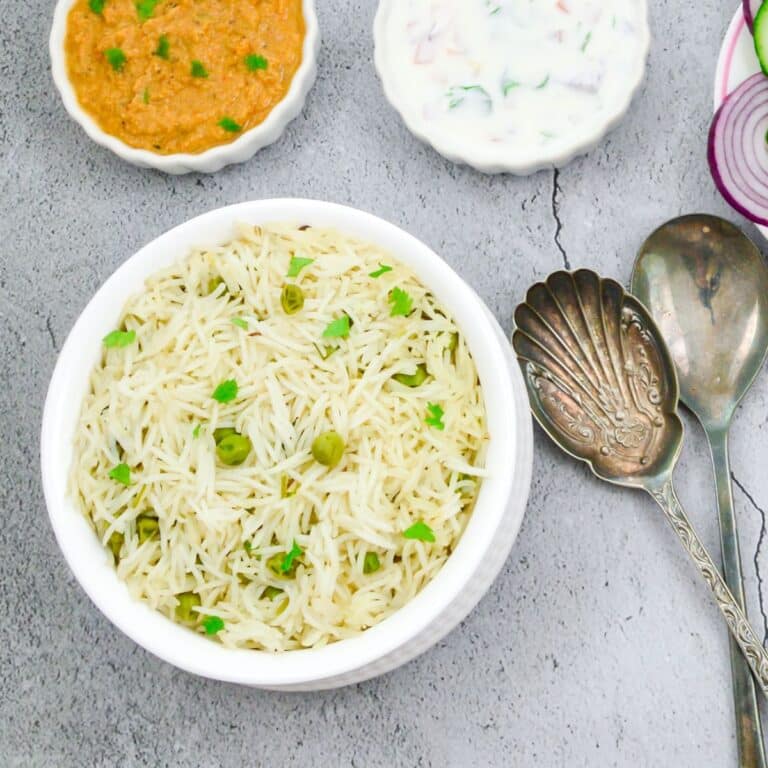 peas pulao in a white bowl and 2 spoons placed on a wooden chopping board along with a bowl of gravy, raita, sliced onion on a granite.