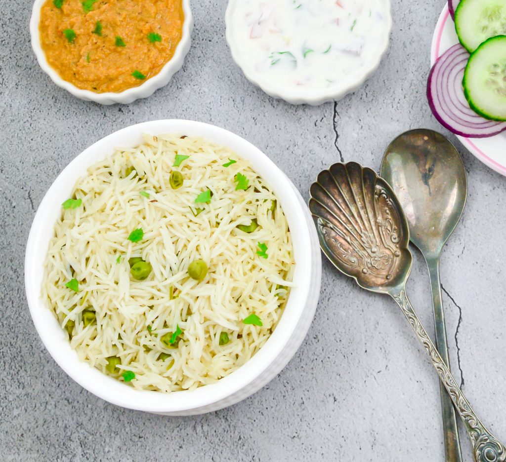 peas pulao in a white bowl placed on a granite along with a bowl of gravy, raita, sliced onion and 2 spoons.