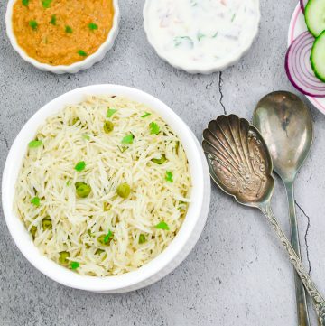 peas pulao in a white bowl placed on a granite along with a bowl of gravy, raita, sliced onion and 2 spoons.