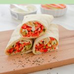 three tortilla wraps filled with vegetables placed on a wooden board with crushed pepper sprinkled on board with cheese tub, bowl of vegetable slices behind and brown text overlay on green background.