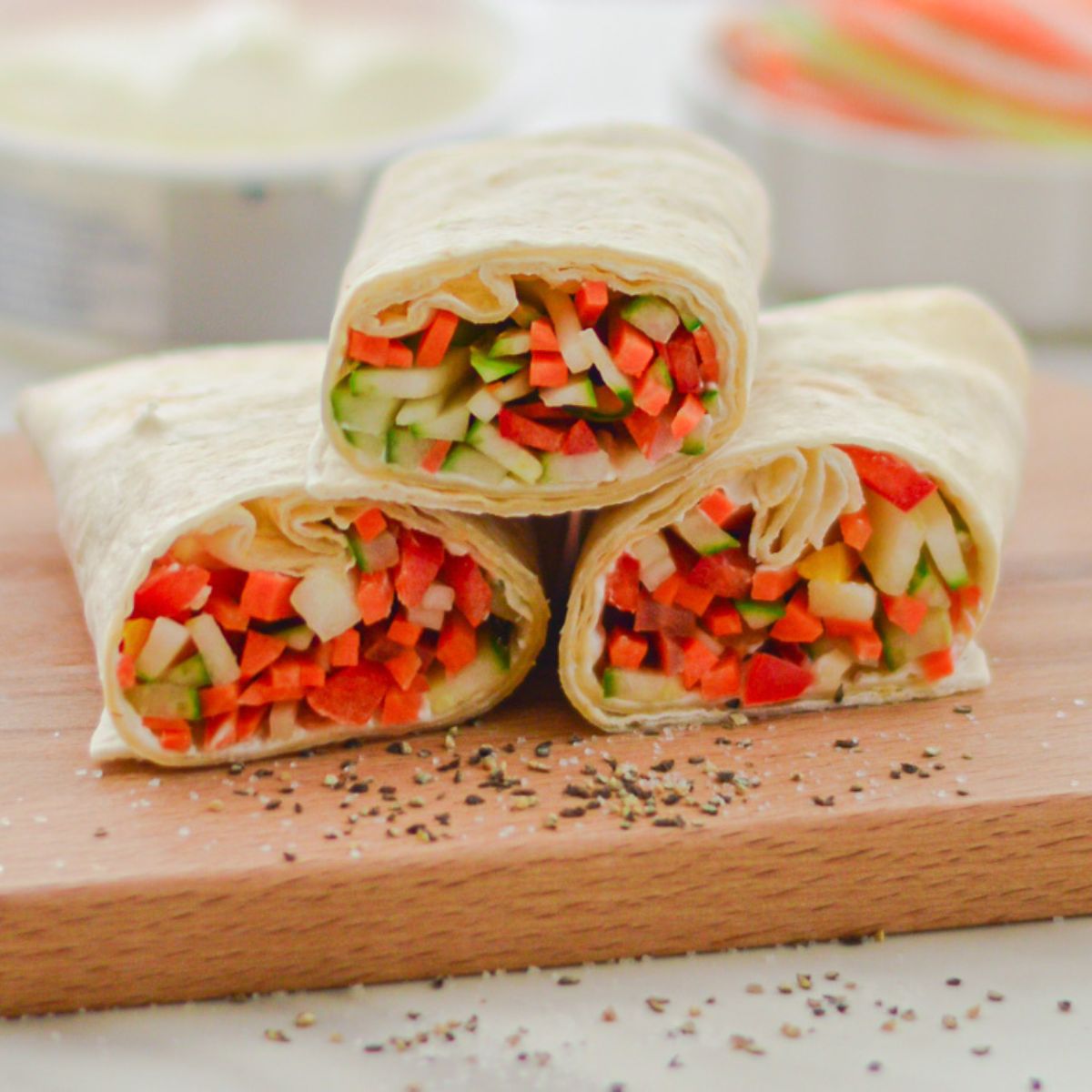 three veggie wraps placed on a wooden board with a cheese tub and a bowl of sliced vegetables behind it.