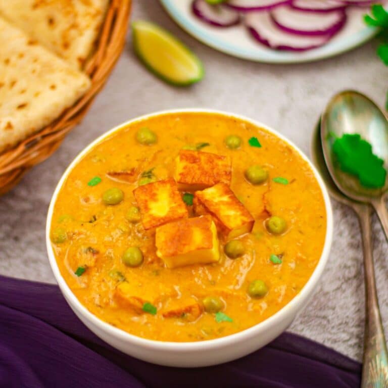 malai matar paneer curry in a white bowl placed on a granite along with a basket of chapatis, a plate of onion rings, bunch of coriander leaves, 2 spoons and a blue cloth.