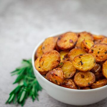white bowl filled with roasted baby potatoes with sprigs of rosemary beside it placed on a grey backdrop.