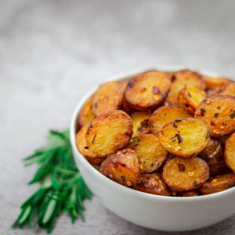 white bowl filled with roasted baby potatoes with sprigs of rosemary beside it placed on a grey backdrop.