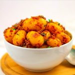 white bowl of bombay aloo placed on a wooden tray with a spoon on side and brown text overlay with green background.