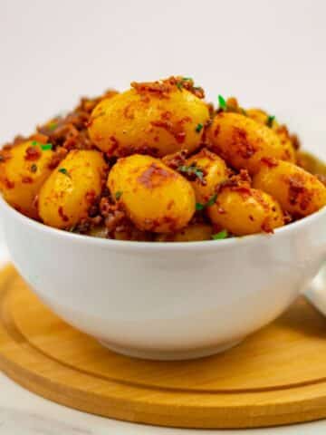 bombay potatoes in a white bowl placed on a round wooden board along with a spoon next to it.