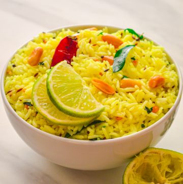 A white bowl of lemon rice topped with 2 half lemon slices, red chilli placed on a marble with a coriander leaves and squeezed lime on the side.