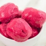 Close up shot of raspberry ice cream scoops in a white bowl placed on marble along with 2 spoons and a cloth.