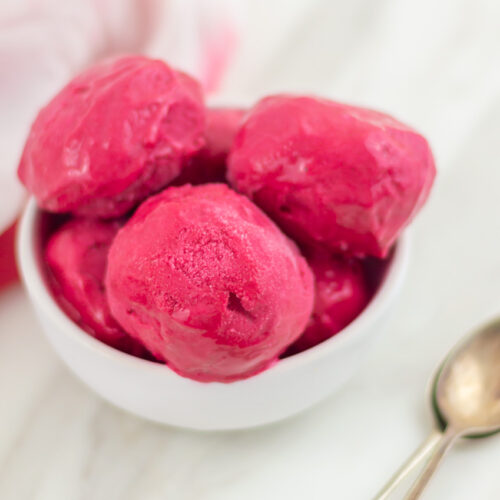 Raspberry ice cream scoops in a white bowl placed on marble along with 2 spoons and a cloth.