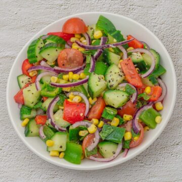 white bowl filled with corn, cucumber, tomato, onion, capsicum salad placed on a granite background.