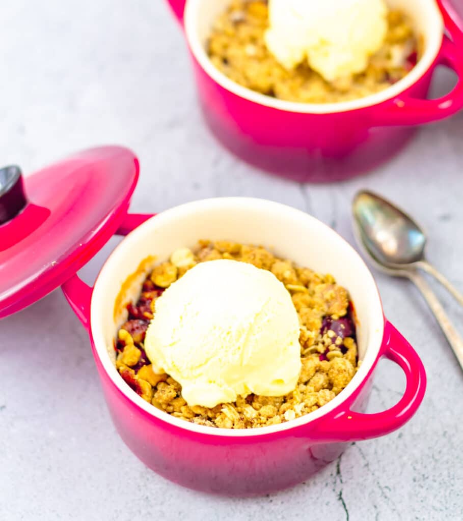 cranberry crisp in two red casseroles topped with a scoop of ice cream and spoons placed on side.